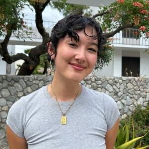 Daisy Okazaki has wavy black hair pulled back and wears hoop earrings and a gray T-shirt. Okazaki stands in front of a stone wall with a tree behind it.